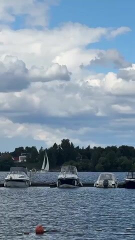 This is a vertical, wide-angle shot of a lake on a cloudy day. The water is a deep blue with ripples. In the foreground, there are several moored motorboats lined up along a wooden dock. The boats are white and have windshields and canopies. Beyond the boats, a lush green forest lines the distant shore. A white sailboat is visible in the distance, its sail catching the light. The sky above is a mix of blue and dramatic, puffy white clouds. There are no visible people, text, or specific events taking place. The scene evokes a peaceful, serene atmosphere typical of a lakeside vacation spot.