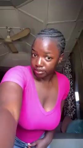 The video shows a dark-skinned young woman standing indoors in front of a camera. She wears a form-fitting, bright pink short-sleeved shirt and a denim miniskirt. The woman has her hair styled in tight braids. She is facing the camera and moves her arms and hands briefly. Behind her, a ceiling fan is visible, along with a light-colored paneled ceiling and a patterned curtain. The room appears small and simply furnished.