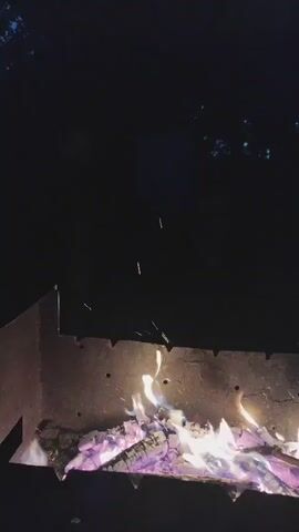 This media file captures a close-up, low-angle view of a bonfire at night, likely in Kolpakovo, Russia. The primary subject is the fire itself, which is burning intensely within a metal brazier or fire pit. The logs are glowing with a vibrant mix of purple and yellow flames, interspersed with bright orange sparks that are actively leaping from the fire. The background is almost entirely dark, suggesting it is nighttime. Sparse, distant lights or stars are faintly visible in the upper portion of the frame, hinting at an outdoor setting, possibly a backyard or a rural area. The overall mood is warm and inviting, evoking the atmosphere of a cozy evening gathering around a fire. No people are clearly visible, though a faint, out-of-focus shape in the upper left corner might be a person's head. The weather appears calm, with no indication of wind. No visible text is present in the media. The focus is entirely on the dynamic and mesmerizing nature of the burning wood and the scattered sparks against the dark backdrop.