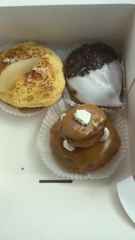 The video opens with a close-up view of a white cardboard box containing three pastries. The pastries are arranged in a triangular formation. The top-left pastry is round and yellowish, topped with a slice of what appears to be pear and dusted with a brown powder. To its right, a second pastry is visible. It is also round, but covered in white icing and dark chocolate sprinkles. The third pastry is located below and to the right of the first two. This pastry is a light brown color, with a caramel-colored glaze and a smaller pastry on top, decorated with white icing.

The camera then pans to the left, revealing a person's arm and shoulder. They are wearing a black top. The person is sitting in a car, as evidenced by the car's interior visible in the background. This includes a dark dashboard, a black circular object, a white travel mug with a lid, a teal colored bottle, and a white mask with pink cloud-like drawings. A person's hand, with visible hair on the forearm, is reaching towards the right side of the frame, presumably towards the box of pastries. The car seat is upholstered in a dark red fabric with white trim.