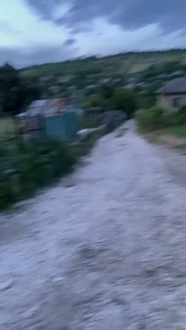 The video captures a moving perspective of a rural setting at dusk. The camera pans across a dirt road leading through a village. On the left side of the road, there are fences and some vegetation. To the right, a house with a light-colored roof is visible, surrounded by grass and some discarded items. Further along the road, trees and other houses can be seen, with a hillside in the background under a cloudy sky. The footage is shaky, suggesting it was recorded while walking or in a moving vehicle.