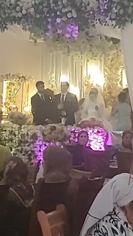 This is a low-resolution video of a wedding reception. The video is shaky and the focus is poor, making it difficult to discern details. In the center of the frame, a bride and groom are seated on a decorated dais. The bride wears a white wedding dress and veil, and the groom is in a suit. Two men in suits stand behind them, possibly officiants or family members. The dais is adorned with white flowers and greenery, and there are floral arrangements in the foreground. A crowd of people are seated in the foreground, mostly out of focus, but some are wearing headscarves. The overall lighting is dim, with some artificial lights illuminating the scene. The video appears to have been shot on a mobile phone.