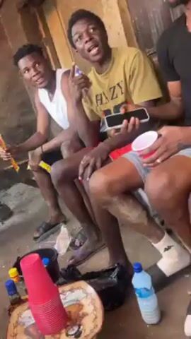 The video shows a group of men sitting outdoors. Several men are sitting on what appears to be a concrete or stone surface. One man is wearing a yellow and black soccer jersey with the 1&1 logo, another is wearing a black t-shirt and grey shorts. Another man is wearing a pink and purple jersey with a logo on the front, and another is wearing a green Adidas t-shirt. Some of the men are holding items, including a red cup and what appears to be a plastic bag. A water bottle is seen on the ground near the men. The background includes a wall and what seems to be a wooden structure. One of the men is seen laughing.