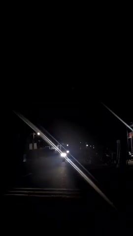 A vehicle with bright headlights approaches on a dark street at night, causing light flares on the lens. Streetlights illuminate the background, where a structure and parked motorcycles are faintly visible. The surroundings are otherwise deeply shadowed.