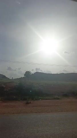 The video shows a sunny rural landscape from a moving vehicle.  The sun is very bright in the sky, obscuring some details. The road is in the foreground. Beyond the road is an area of brown dirt and green vegetation. A hill or low mountain is visible in the background, along with scattered trees.  Power lines run across the sky.