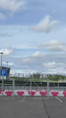 The video shows an empty parking lot with a metal gate in the background. The gate is closed and has a white and pink striped barrier in front of it. There are parking spaces marked on the asphalt in front of the gate. To the left of the gate, there is a blue parking sign with a white "P" and a wheelchair symbol. In the background, there are trees and flags. The sky is blue with white clouds.