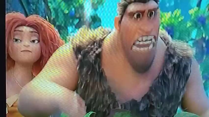 The video shows a scene from an animated movie with two characters, a male and a female, in a lush, green, jungle-like environment. The male character, who is larger and has spiky brown hair, is in the foreground, looking to his left with his mouth open in an expression of surprise or alarm. He is wearing a fur vest. The female character, who has bright orange, curly hair and is wearing a yellow garment, is to his left and slightly behind him. She is looking down with a somber expression. The background is filled with large green leaves and blue and pink flowers. Text overlays are visible at the top of the screen, reading "The Croods: A New Age" and what appears to be Arabic subtitles. The lighting is bright and suggests it is daytime.