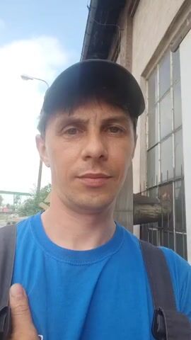 The video shows a man wearing a blue t-shirt and a black baseball cap. He is standing outside, possibly at a construction site or industrial area, as indicated by the background which includes a building with a window and some scaffolding or construction materials. The man is looking directly at the camera, and the video appears to be a selfie. The lighting suggests it is daytime, and the sky is visible in the background.
