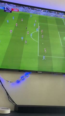 The video shows a Thomson TV displaying a soccer game between Manchester City and Arsenal. The score is 1-0 in favor of Manchester City at the 16:17 mark of the game. Players wearing light blue uniforms (Manchester City) and red and white uniforms (Arsenal) are positioned on a green soccer field.  A string of blue LED lights is attached to the bottom of the TV.  A black sound bar and a white Apple TV are visible below the TV.