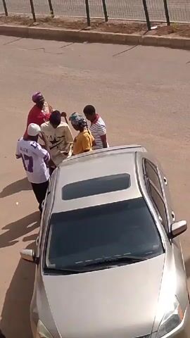 Four people are gathered near a silver sedan with its sunroof open. One person, wearing a pink headscarf and a red shawl, is holding a walking stick and appears to be interacting with the others. The setting appears to be a street or parking area with a sidewalk and a fence in the background. The lighting suggests it is daytime.