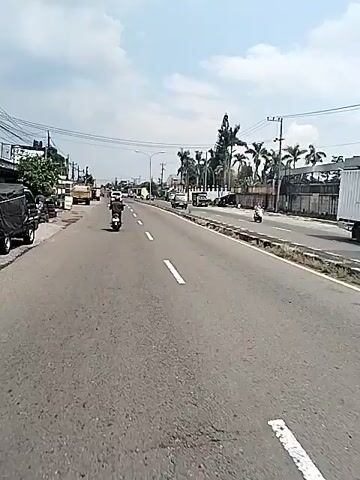 The video shows a person riding a scooter on a road, from the perspective of someone else on a scooter. The rider is wearing a dark shirt and a red helmet. The road has white lane markings. There are other vehicles on the road including cars and trucks. The sky is partly cloudy. There are power lines and light poles visible. There are palm trees and other trees in the background. On the side of the road, there are piles of tires and buildings.