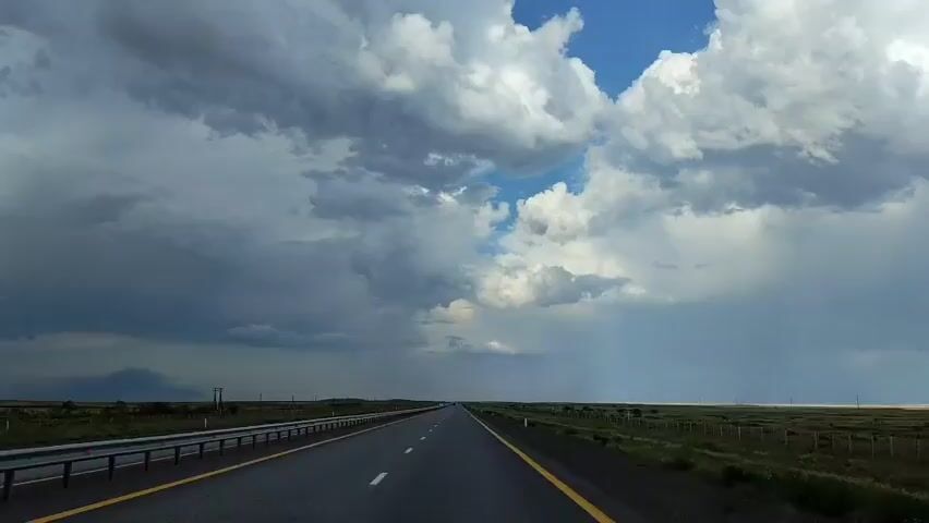 The video shows a forward-facing view from a vehicle driving on a highway. The road has two lanes in each direction separated by a yellow line and white dashed lines. On the left side of the road, there is a metal guardrail and a grassy shoulder. On the right side, there is a fence and more grassy terrain. The sky is filled with dramatic, dark storm clouds, with patches of blue sky visible in the distance. There is a visible rain shower in the distance to the right, indicated by a grey curtain of rain falling from the clouds. The overall environment appears to be open, rural land. There are no people or other vehicles visible in the immediate frame.