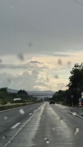 This is a vertical video taken from inside a vehicle, looking forward through a wet windshield. The road ahead is wet and reflective, suggesting recent rain. There is one car visible in the distance on the right lane, moving away from the camera. A bridge-like structure spans the highway in the mid-distance. The sky is cloudy with some lighter areas, possibly indicating the sun breaking through. Trees and foliage line the right side of the road.
