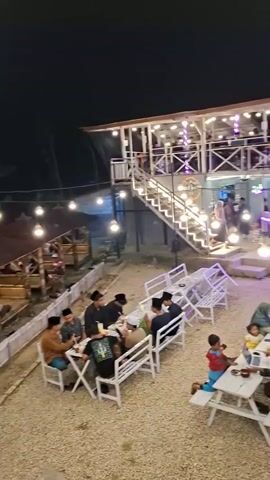 The video shows an outdoor dining area at night. Several picnic tables with benches are set up on a gravel surface, illuminated by string lights. People are seated at some of the tables, eating and socializing. In the background, a two-story building with an open-air second floor is visible, also lit with string lights and with people inside. A staircase leads up to the second floor. To the right of the building, several motorcycles are parked. Further in the background, there are more lights and what appears to be a road with a small building or stall on the side. Some palm trees are also present in the foreground. The overall atmosphere appears to be casual and lively.