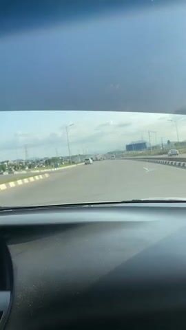 A person is driving on a highway. The car is moving forward, and other vehicles can be seen in the distance. The road is divided by lane markings, and streetlights line the sides. Music is playing in the car. The sky is visible through the windshield, with clouds and some haze.