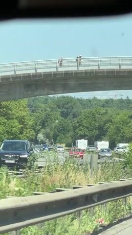The video begins showing the underside of a bridge with two figures standing on the bridge above. The camera then pans down to reveal a highway below the bridge, with cars and RVs traveling or parked along the side of the road. Lush green trees and vegetation surround the highway. The sky is clear and blue. The video appears to be taken from a moving vehicle.