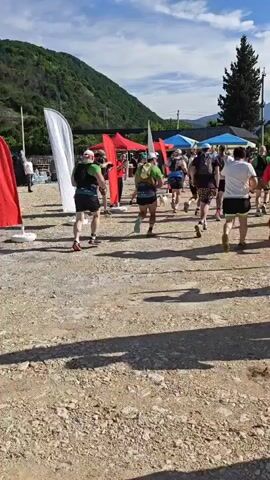 A group of people wearing athletic attire are running away from the camera on a dirt and gravel path, with other people standing on the side, some cheering. In the background, there are trees, buildings with red and blue awnings, power lines, and mountains under a blue sky with scattered clouds.