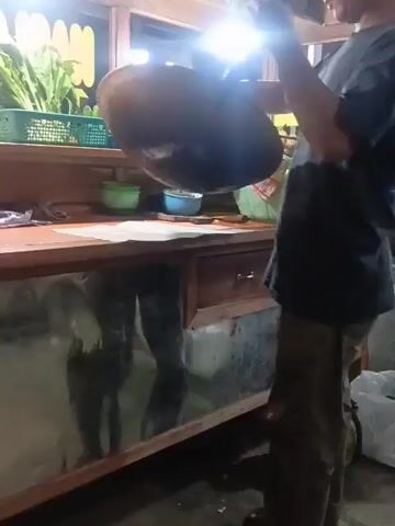 A person, wearing a dark t-shirt and tan pants, stands by a wooden counter. They are holding a large, dark wok, tilted downwards, as if to empty its contents onto a white paper placed on the counter. The wok appears to be made of metal and shows signs of use. The counter has a glass front, through which a reflection of what appears to be legs and a torso is visible, suggesting another person or the same person from a different angle. To the left of the counter, there is a green plastic crate filled with leafy vegetables. In the background, there are blurred lights and structures that indicate an outdoor setting, possibly a food stall at night. The overall scene depicts food preparation or serving.