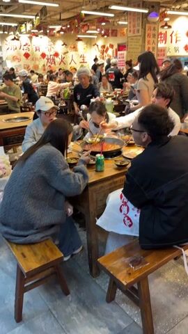 The video shows a bustling restaurant with numerous customers seated at tables. A group of six people are gathered around a square wooden table with a hot pot in the center, which appears to be a tomato-based broth. A young woman in a white apron is serving food from the hot pot with a ladle, and others are eating from bowls with chopsticks. On the table are several small dishes, a can of drink, and serving utensils. The customers are dressed in casual attire, and there's a white plastic bag with red printing near one of the seated men. The restaurant is decorated with Chinese lanterns and wall hangings with Chinese characters. Wait staff are seen moving between tables, attending to customers. The environment is noisy and lively.
