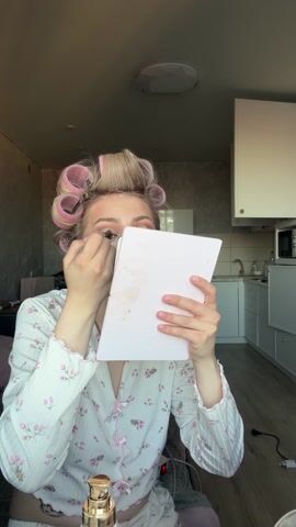 A young woman is doing her makeup. She is wearing a white floral pajama set, and her hair is in rollers. She is holding a small mirror in her left hand and applying eyeliner with a brush in her right hand. She smiles at the camera. The background shows a small apartment with a kitchen area, including cabinets, a sink, and a microwave. The lighting is soft and natural.