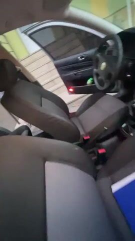 The video begins with a blurry interior view of a car's passenger seat and dashboard, a blue binder resting on the seat. The scene then shifts to an exterior shot of a silver station wagon with its rear passenger door open. A young person in a black t-shirt and shorts is leaning into the open rear of the car, holding up two fingers in a peace sign gesture, facing the camera. They appear to be in a playful mood. In the background, a yellow and green building is visible with a few other people standing near a railing. The car is parked on an asphalt surface. The video ends abruptly.