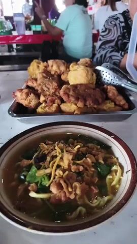 The video is a close-up, POV shot of food. In the foreground, a bowl of noodles with broth, greens, and fried shallots is visible. A hand is holding up a piece of golden-brown fried food, possibly a fritter or fried tofu, over the bowl. In the background, a tray of similar fried items is on a table, and blurred figures of people are seated at tables in what appears to be an outdoor food setting. The lighting is natural and seems to be daytime.