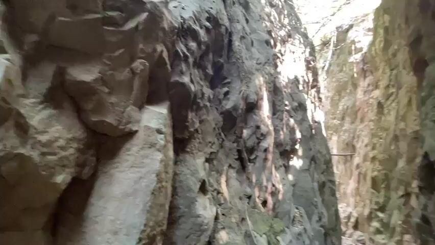 The camera moves through a narrow passage with high rocky walls on both sides.  The rocks are weathered and vary in color from light gray to dark brown, some with visible moss. The passage floor is a dirt path with small rocks and vegetation. Sunlight filters through the top, illuminating the path ahead. The atmosphere is still and quiet with the exception of sounds of cicadas.