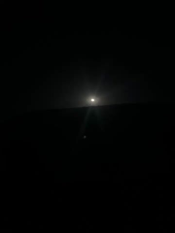 The video shows a bright moon in a dark sky, positioned above a silhouette of a hill or trees.  There is a faint light streak emanating downwards from the moon.  In the foreground, a faint green lens flare is visible. The sound of a dog barking can be heard.