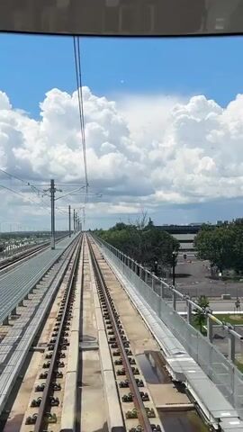 The video shows a view from a train moving along elevated tracks on a bright, cloudy day. The train tracks run parallel on the left side, with a metal railing on the right side. Overhead power lines are visible above the tracks. In the distance, trees and buildings can be seen. The sky is blue with large, fluffy white clouds. The camera appears to be positioned at the front of the train, looking forward along the tracks.