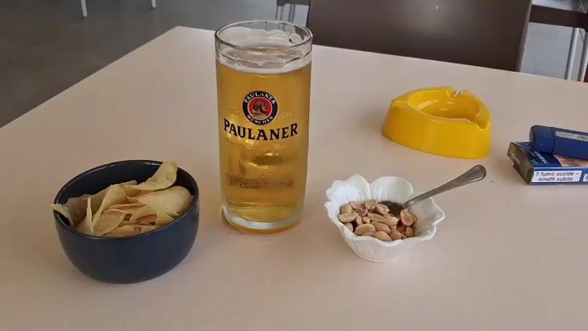 The video opens with a close-up shot of a table, likely on a balcony or terrace in Savona, Italy. On the tabletop, a tall glass of Paulaner beer with a foamy head is central, featuring the brand's logo and "MÜNCHEN 1634" text. To its left, a dark grey bowl is filled with potato chips, and to its right, a white, flower-shaped bowl holds roasted peanuts along with a small spoon. A yellow ashtray and a blue-and-white cigarette packet, bearing Italian warnings "Il fumo uccide" (smoking kills) and "smetti subito" (quit immediately), complete the arrangement, suggesting a leisure activity. The beer glass is frosted with condensation, indicating it is cold.

The camera then pans to reveal the view from the vantage point. A red planter box and a black backpack are briefly visible as the perspective shifts towards the beach and the sea. The scene captures a wide expanse of the sea under a slightly overcast but bright daytime sky. Gentle waves break upon the sandy shore, where several closed red and white beach umbrellas and some vertical poles are present. Further out in the water, a line of yellow buoys marks an area, and at least two individuals can be seen on an inflatable raft or paddleboat, enjoying the water. The overall impression is one of a relaxing day at the Italian coast.