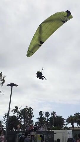 A tandem paragliding flight is in progress against a cloudy sky. The paraglider, which is lime green with black accents, is positioned high and to the right of the frame. Below the paraglider, two people are suspended in a harness, with the person in front appearing to be the pilot and the person behind a passenger. They are seated facing forward, with their legs extended. The paraglider is attached to the harness by numerous lines. In the background, a row of palm trees and other foliage is visible. The scene appears to be near a coastal area, with some buildings and a street in the foreground. Several people are gathered on the ground, some looking up at the paraglider. One person on the ground is holding up a phone, likely to take a photo or video. The overall impression is that of a recreational activity taking place in a public outdoor space.