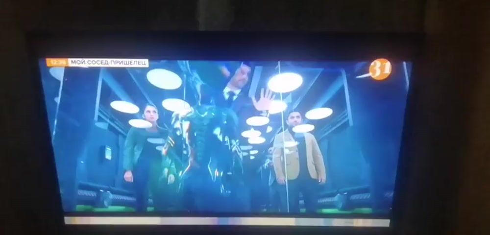 The video displays a television screen showing a scene from a show or movie. The scene appears to be set in a futuristic or industrial environment with blue lighting and circular ceiling lights. In the foreground, several people are visible. On the left, a woman in a green jacket and dark pants stands next to a man in a suit. In the center, a man in a suit and tie stands facing away from the camera, holding his hand up as if to stop someone. To the right, another man in a suit and tie is visible. In the background, there are several individuals in what appear to be security uniforms, some holding weapons. There is also a large, metallic, possibly robotic figure in the center of the scene, glowing with blue light. Text overlays on the screen indicate a time stamp of 12:36 and the title "МОЙ СОСЕД-ПРИШЕЛЕЦ" (My Neighbor is an Alien), along with a channel logo featuring the number 31. The overall impression is a dramatic or action-oriented scene involving multiple characters and possibly a conflict.