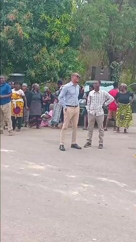 The video depicts a daytime protest or demonstration primarily involving women, with some men and children also present, on an asphalt road in what appears to be Ungwan Tanko, Nigeria. The scene transitions from a tree-lined area near some parked vehicles and a small building to a more open road with parked cars and motorcycles, and distant industrial structures, possibly part of a refinery. A sign on a building reads "THE NIGERIA POLICE FORCE NNPC OUT-POST KADUNA," indicating the location's connection to the Nigerian National Petroleum Corporation.

The participants are engaged in an active protest, with several women holding up handmade signs and some appearing to shout or chant. One woman is seen sitting on the ground in a posture of protest. Visible text on the signs includes demands such as:
*   "PAY US OUR CROPS!!"
*   "HUNGER IS KILLING US"
*   "PAY US OUR DAMAGE"
*   "NO FOOD NO WORK"
*   "After Taking Our Record We Have Not Hard [sic] From You Again!!!"
*   Another sign is partially visible, reading "WHAT DO YOU WANT..."

Many women are holding green leafy branches, possibly as a symbol of their land or crops. The mood among the protesters appears to be one of frustration, anger, and desperation, as conveyed by their shouting and the content of their signs. Some individuals are seen observing the protest, including a man recording with his phone. The overall setting suggests a community expressing grievances, likely concerning issues related to agricultural land, damages, and economic hardship, directed towards the entities represented by the nearby facilities.