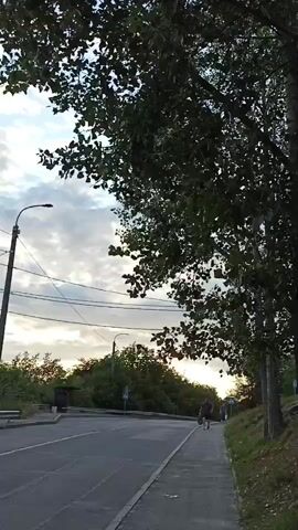 A person is walking on a sidewalk next to a road under a cloudy sky. There are trees and power lines in the scene.
