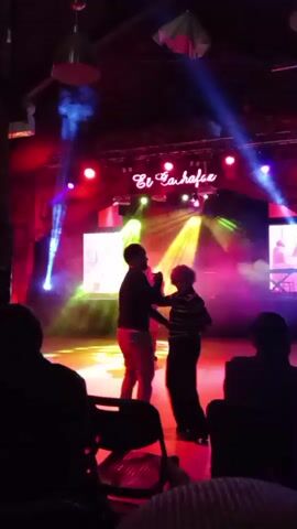 This is a video taken from an audience's perspective in a dimly lit club. A couple is dancing on a stage illuminated by colorful spotlights and stage lighting. The stage backdrop has the words "El Zahara" in cursive neon lights. Other people are visible in the background, some dancing and some seated. The focus is on the dancing couple.