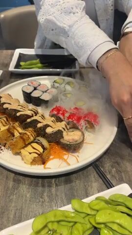 The video shows a table at a sushi restaurant. A white plate of sushi is in the center of the frame. The sushi includes several types of rolls. Some rolls are topped with pink roe and avocado. Other rolls are fried and drizzled with a creamy sauce. A person wearing a white lace shirt is visible on the right side of the frame, reaching towards the sushi with chopsticks. There is a dish of edamame in the foreground. A small square plate with edamame is also visible in the background. The table is dark wood.