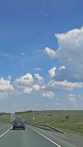 A black SUV drives down a highway through a green, rural landscape under a bright blue sky with white clouds. The camera is positioned behind and slightly above the SUV, capturing the road ahead and the surrounding scenery.