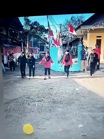 The video shows a group of five people walking down a dirt road, blindfolded. They are all wearing casual clothing and appear to be participating in a game or activity. There are houses and trees in the background, as well as Indonesian flags, suggesting a festive or celebratory occasion. A yellow balloon is visible on the ground in the foreground. The participants are moving in a somewhat disorganized manner, with some appearing to stumble or veer off course. The overall atmosphere seems lighthearted and playful.