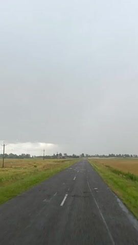 A point of view shot from inside a vehicle shows a long, narrow, asphalt road stretching into the distance. The sky is overcast and gray, with hints of clouds on the horizon. On either side of the road are fields of grass and crops, with some scattered buildings and trees visible in the far distance. The road itself appears to be in poor condition, with several potholes and patches of standing water visible. A windshield wiper is visible in the foreground, and it appears to be in motion, suggesting that it is raining or that water is being sprayed onto the windshield. The overall impression is one of a desolate and somewhat melancholic rural landscape.