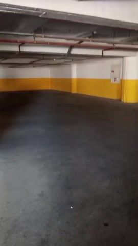The video shows a first-person perspective of walking through an underground parking garage. The garage has concrete floors and ceilings, with white support columns painted with a yellow band near the bottom. Orange pipes are visible along the ceiling. In the background, several cars are parked, including a white car and a dark-colored SUV. There are also yellow and white markings on the walls. The camera moves smoothly, suggesting a person is walking at a moderate pace. The lighting is typical of an indoor parking facility. There is a small office or security booth visible in the distance at one point, with a person inside.