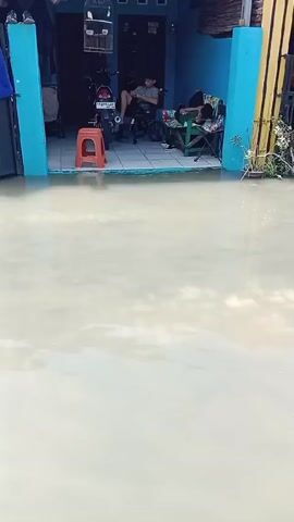 The video shows a flooded residential street. The water level is high enough to cover the bottom of the house and the street in front of it. In front of one of the houses, which is painted blue and yellow, a person is sitting in a chair and looking at a phone, and another person is lying on a chair asleep. A motorcycle is parked behind them, along with an orange stool. The houses are situated next to each other and are all flooded to a similar degree. The water is murky and brown. Some plants are partially submerged near the house. The video is taken from a slightly elevated position.