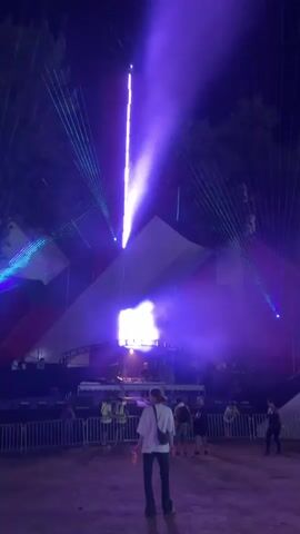A video clip shows a group of people at an outdoor night concert. The stage features bright purple and blue lasers. In the foreground, a person wearing a white shirt and dark pants stands facing away from the camera, holding a clear bottle with a yellow cap. Several other people are visible in the background, some near the stage and others further back. The ground appears to be dirt or sand, and there is a fence separating the crowd from the performance area. The overall atmosphere is that of a lively event with vibrant lighting effects.