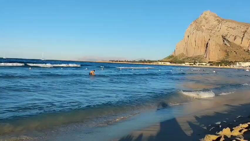 The video shows a beach scene with the ocean and the sky. The water is blue with waves and there are some boats visible in the distance. A man is seen wading in the water, approaching the shore. The sun appears to be setting or rising, casting a warm light on the scene. The beach itself is sandy.