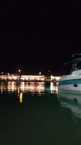 A nighttime view from a body of water shows a brightly lit building in the background with reflections on the water's surface. In the foreground, the rear section of a white and teal boat is visible, partially submerged in the dark green water. The sky is black.