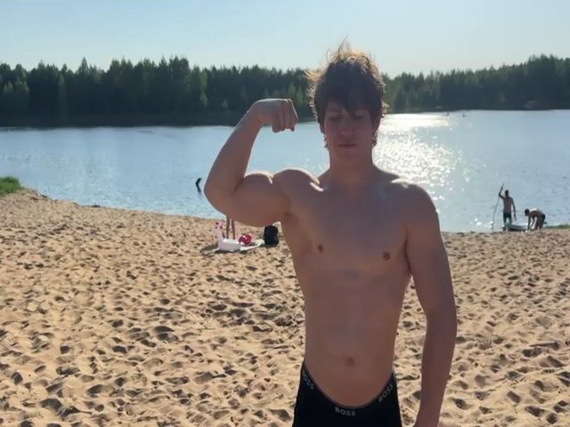 A young man stands shirtless on a sandy beach, flexing his left bicep. He is wearing dark boxer briefs with the word "BOSS" on the waistband. Behind him is a lake with a line of trees on the opposite shore. There are a few people in the water and on the beach in the background, including a person on a paddleboard. A small inflatable flamingo is visible on the sand behind the man. The sky is clear and blue, indicating a sunny day.