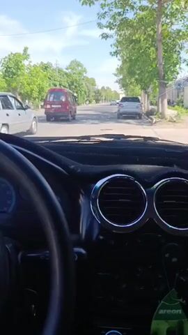 The view from inside a car shows a road lined with trees and buildings. Several other vehicles are visible ahead on the street. A rearview mirror and parts of the car’s dashboard are visible inside the car. The sky is blue with scattered clouds.
