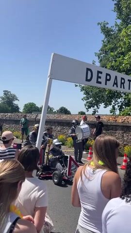 A group of people are gathered outdoors on a sunny day, with a large sign that reads "DEPART" overhead. In the center, a go-kart with two people in it is being pushed forward by another person. One person in the go-kart is wearing a helmet and a gray jumpsuit. Another person is playing a large drum. In the background, there are trees, a stone wall, and a building that resembles a castle. A large screen is visible, showing a crowd of people. The event appears to be a race or a parade, with spectators lining the route.
