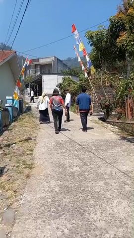 A group of five people are walking on a paved road in a rural area. The road is lined with grass and small bushes. There are houses and trees on either side of the road. Several colorful flags are strung across the road on poles. The weather is sunny and clear.