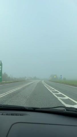 The video shows a driver's point of view from inside a car driving on a straight, wide, paved road. The road has white lane markings and several overhead streetlights. The surrounding environment is enveloped in thick fog, obscuring the view beyond the immediate roadside. Road signs are visible, although their details are slightly obscured by the fog.