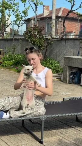 Here's a description of the video:

A young woman is sitting on a grey folding chaise lounge on a sunny day, holding a small, light-colored dog in her lap. She is wearing a white crop top, patterned pants, and blue shoes, with sunglasses pushed up on top of her head. She is gently petting and talking to the dog.  The dog is mostly white with some darker markings around its face. 

The setting appears to be a backyard or patio area. The ground is paved with stone or concrete slabs. Behind the woman is a stone or concrete fence and some greenery. In the background, there is a brick house with a red tile roof.